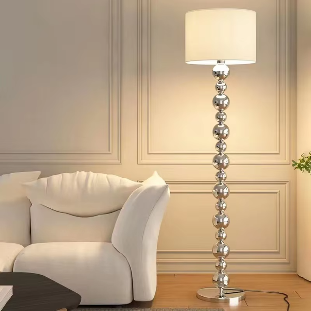 This is a modern light luxury-style silver spherical decorative floor lamp, with a silver metal pole composed of multiple spherical elements connected in series, paired with a beige fabric lampshade. It features a minimalist and textured design, with soft and warm lighting. It is used for space lighting and decoration, creating a light luxury and warm atmosphere. Suitable for modern light luxury living rooms, bedroom corners, high-end apartments and other places.
