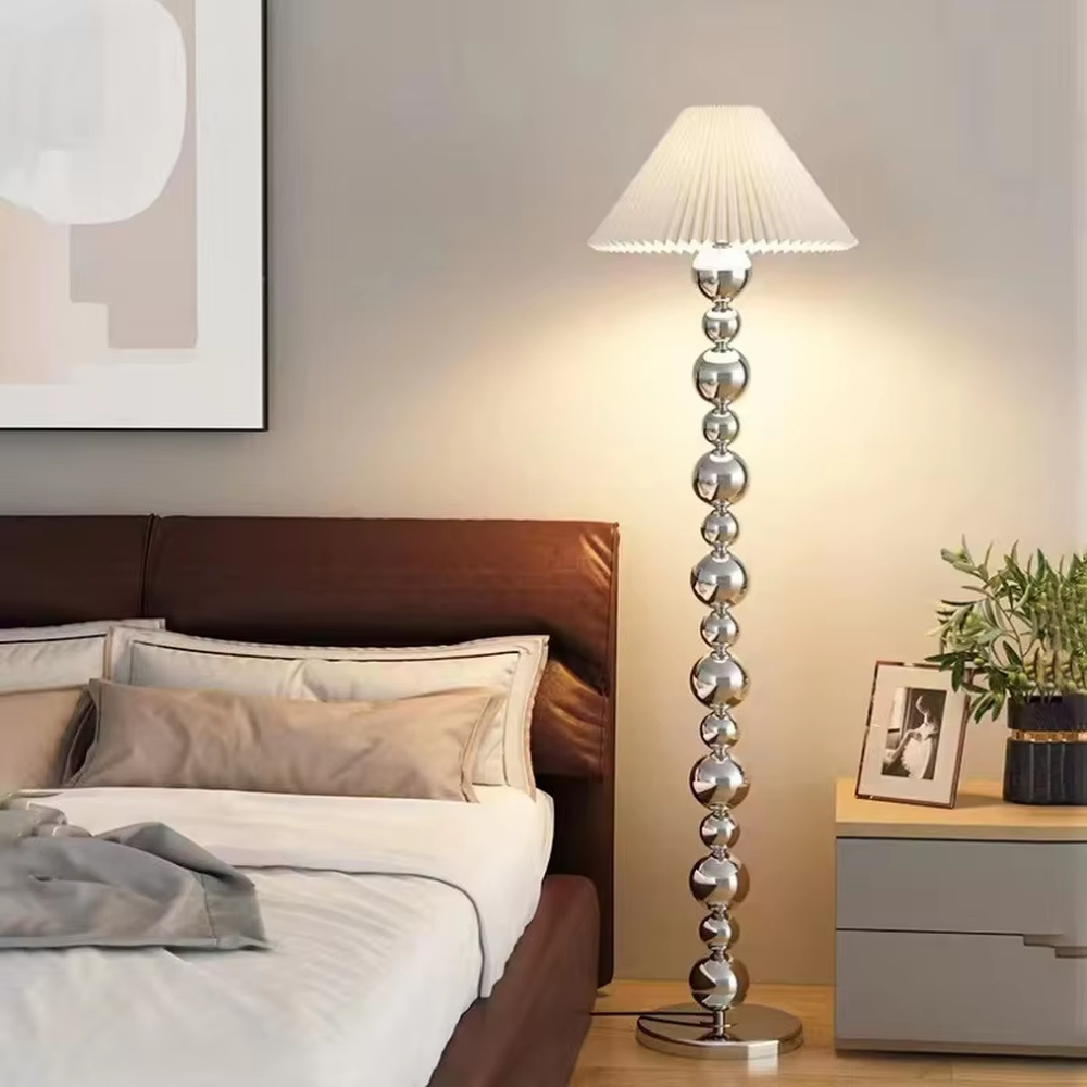 This is a light luxury exquisite silver metal ball column floor lamp, with silver metal balls layered to form a column, paired with a beige fabric round lampshade. It features a light luxury and exquisite design, with soft lighting. It is used for space ambient lighting and decoration, creating a light luxury elegant atmosphere. Suitable for light luxury-style living rooms, modern minimalist bedrooms, high-end hotel guest rooms and other places.