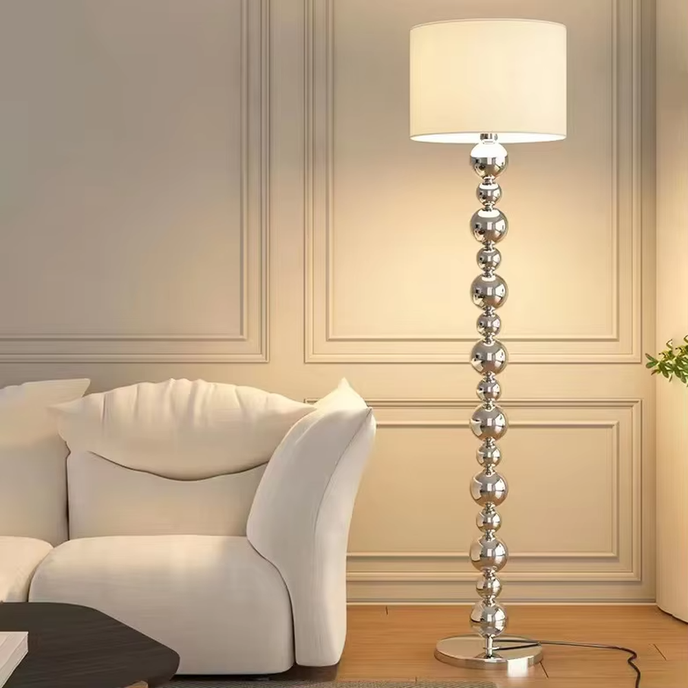 This is a light luxury exquisite silver metal ball column floor lamp, with silver metal balls layered to form a column, paired with a beige fabric round lampshade. It features a light luxury and exquisite design, with soft lighting. It is used for space ambient lighting and decoration, creating a light luxury elegant atmosphere. Suitable for light luxury-style living rooms, modern minimalist bedrooms, high-end hotel guest rooms and other places.