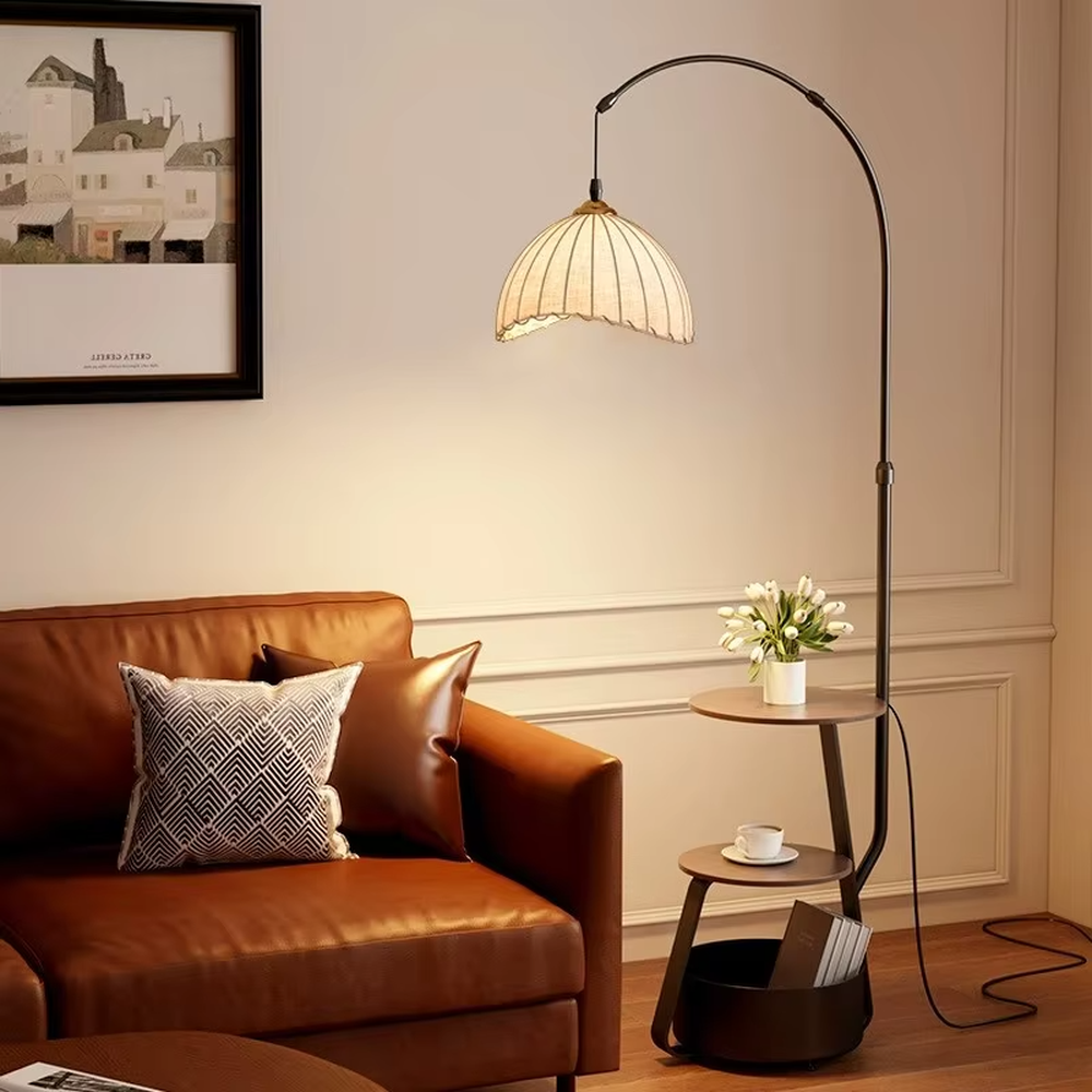 This is a modern multi-functional arc floor lamp with a black metal arc pole, a shell-shaped fabric lampshade, and integrated multi-layer storage shelves, featuring a minimalist and practical design. It is used for space lighting, storage, and decoration, creating a warm and practical atmosphere. It is suitable for modern home living rooms, small-sized residences, leisure reading areas and other places.