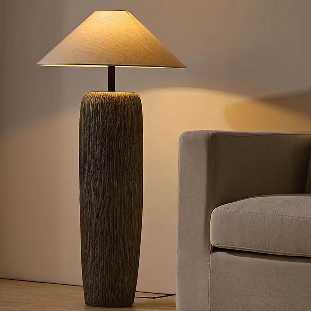 This is a natural wabi-sabi style gray textured column floor lamp, with a gray textured column base, a beige fabric conical lampshade, and a black metal pole connection. It features a minimalist and natural design with a rustic texture. It is used for space lighting and decoration, creating a warm and rustic atmosphere. Suitable for wabi-sabi style living rooms, Japanese-style bedrooms, natural-style homestays and other places.