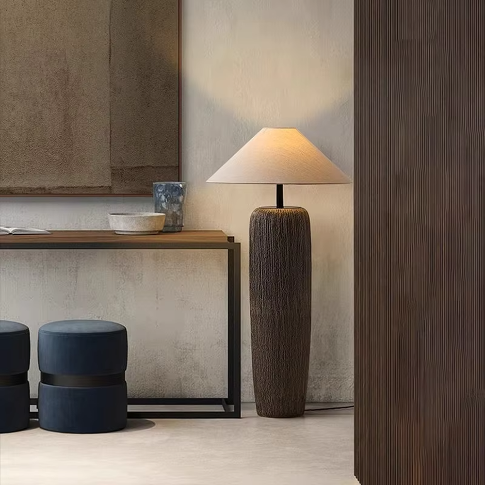 This is a natural wabi-sabi style gray textured column floor lamp, with a gray textured column base, a beige fabric conical lampshade, and a black metal pole connection. It features a minimalist and natural design with a rustic texture. It is used for space lighting and decoration, creating a warm and rustic atmosphere. Suitable for wabi-sabi style living rooms, Japanese-style bedrooms, natural-style homestays and other places.