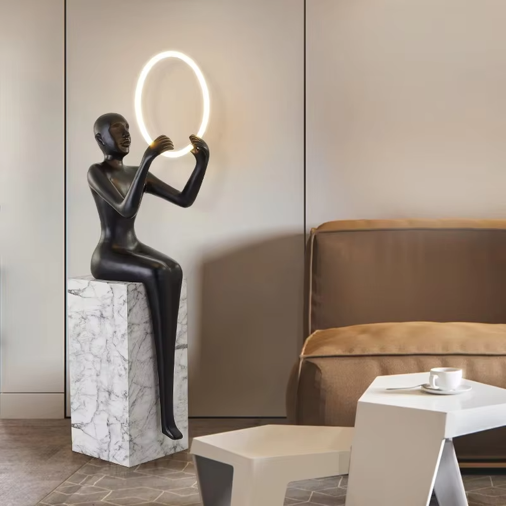 This is an artistic installation-style human sculpture ring floor lamp, featuring a black human sculpture shape paired with a circular LED light. It has a strong sense of art and avant-garde, with soft lighting and visual impact. It is used for space lighting and artistic decoration, creating a modern artistic and avant-garde atmosphere. Suitable for art exhibition halls, high-end villa living rooms, designer studios and other places.