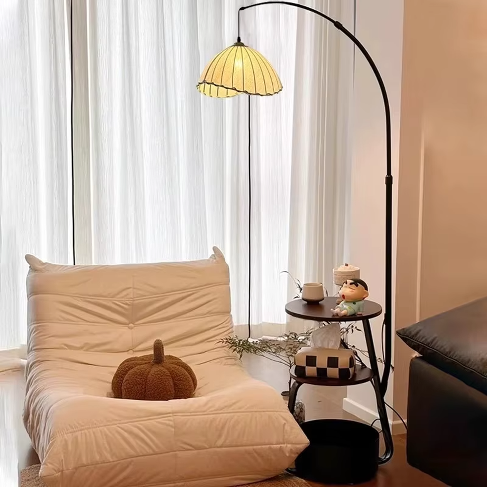 This is a modern multi-functional arc floor lamp with a black metal arc pole, a shell-shaped fabric lampshade, and integrated multi-layer storage shelves, featuring a minimalist and practical design. It is used for space lighting, storage, and decoration, creating a warm and practical atmosphere. It is suitable for modern home living rooms, small-sized residences, leisure reading areas and other places.