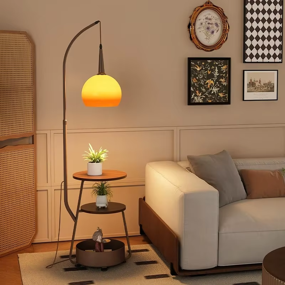 This is a modern multi-functional floor lamp with a curved metal pole, a gradient orange-yellow spherical lampshade, and integrated multi-layer storage shelves, featuring a minimalist and practical design. It is used for space lighting, storage, and decoration, creating a warm and practical atmosphere. It is suitable for modern home living rooms, small-sized residences, creative studios and other places.