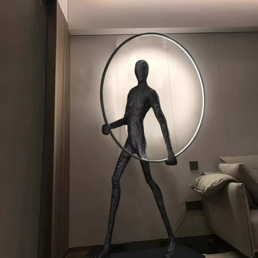 This is an artistic installation-style human sculpture ring floor lamp, featuring a black human sculpture shape paired with a circular LED light. It has a strong sense of art and avant-garde, with soft lighting and visual impact. It is used for space lighting and artistic decoration, creating a modern artistic and avant-garde atmosphere. Suitable for art exhibition halls, high-end villa living rooms, designer studios and other places.