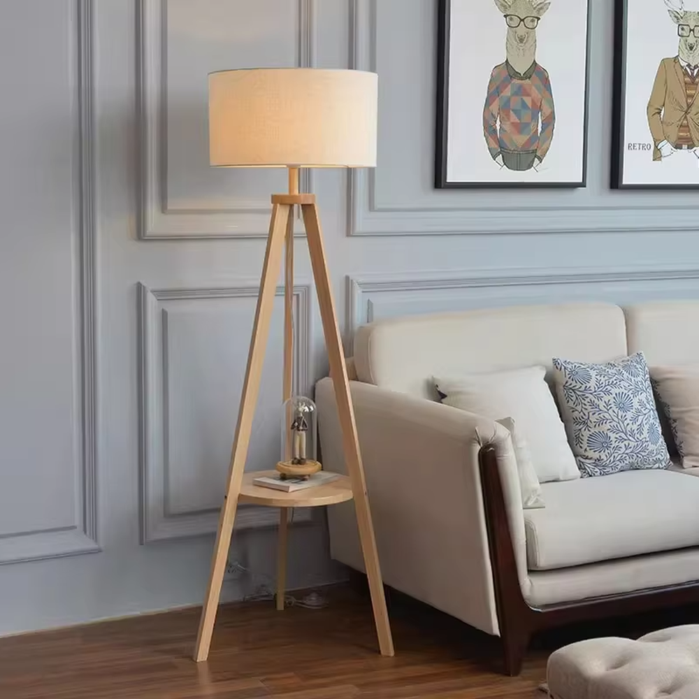 This is a Nordic-style wooden tripod floor lamp, with a light wood tripod stand (with a circular storage table) and a beige fabric cylindrical lampshade, featuring a minimalist and natural design with a warm ambiance. It is used for space lighting and storage decoration, creating a warm and natural atmosphere. It is suitable for Nordic-style home living rooms, minimalist residences, leisure reading areas and other places.