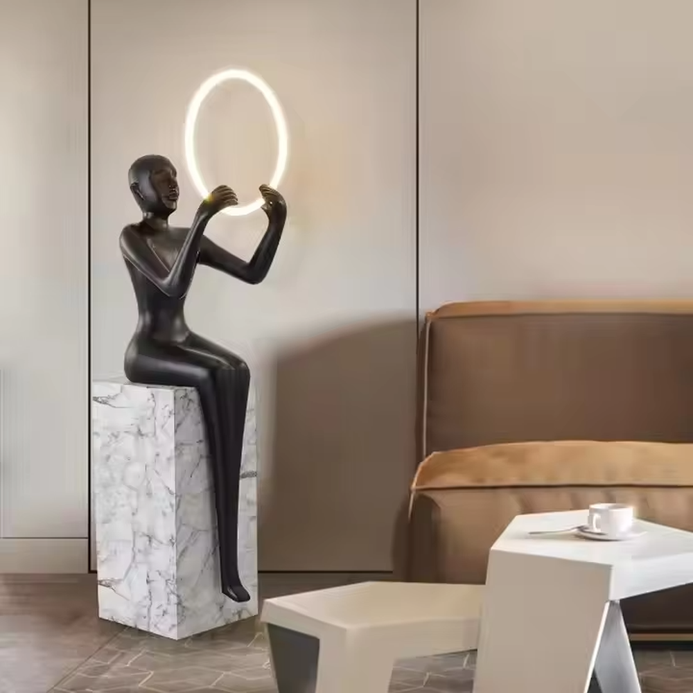 This is an artistic installation-style figure sculpture floor lamp, with a black figure sculpture sitting on a white marble base, holding a circular luminous lamp with both hands, featuring strong artistic sense. It is used for space lighting and artistic decoration, creating a modern artistic atmosphere. Suitable for art exhibition halls, high-end hotel lobbies, corners of modern art home living rooms and other places.