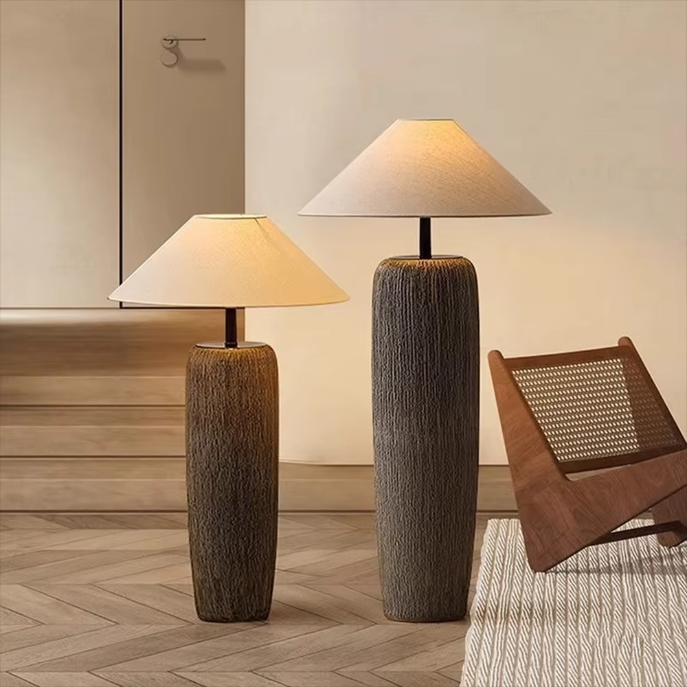 This is a natural wabi-sabi style gray textured column floor lamp, with a gray textured column base, a beige fabric conical lampshade, and a black metal pole connection. It features a minimalist and natural design with a rustic texture. It is used for space lighting and decoration, creating a warm and rustic atmosphere. Suitable for wabi-sabi style living rooms, Japanese-style bedrooms, natural-style homestays and other places.