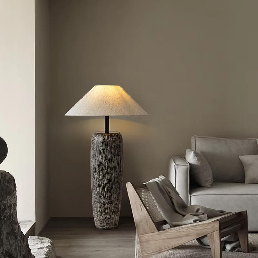 This is a natural wabi-sabi style gray textured column floor lamp, with a gray textured column base, a beige fabric conical lampshade, and a black metal pole connection. It features a minimalist and natural design with a rustic texture. It is used for space lighting and decoration, creating a warm and rustic atmosphere. Suitable for wabi-sabi style living rooms, Japanese-style bedrooms, natural-style homestays and other places.