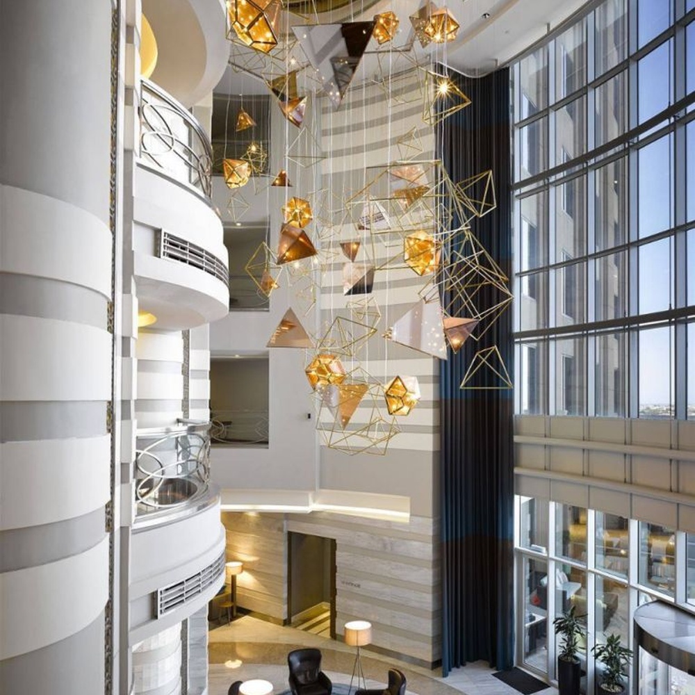 This modern geometric art chandelier is composed of intertwined gold metal lines forming an abstract frame, paired with geometric-shaped liuli/acrylic ornaments. Light refracts through them to emit a brilliant halo, serving both lighting and installation art purposes, suitable for high-ceiling commercial spaces such as hotel lobbies and high-end clubs.