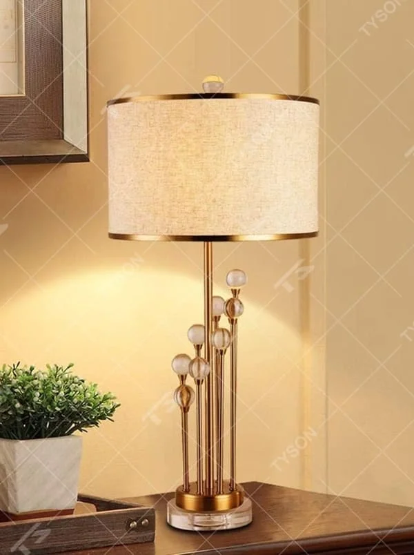 This is a modern luxury-style table lamp with a golden metal stand, multiple spherical decorations, and a beige fabric lampshade that transmits light softly. It has an exquisite and layered design. It is used for local desktop lighting and decoration, combining practicality and aesthetics. It is suitable for modern home living room side tables, bedroom bedside tables, as well as high-end hotel guest rooms, luxury cafes and other places.