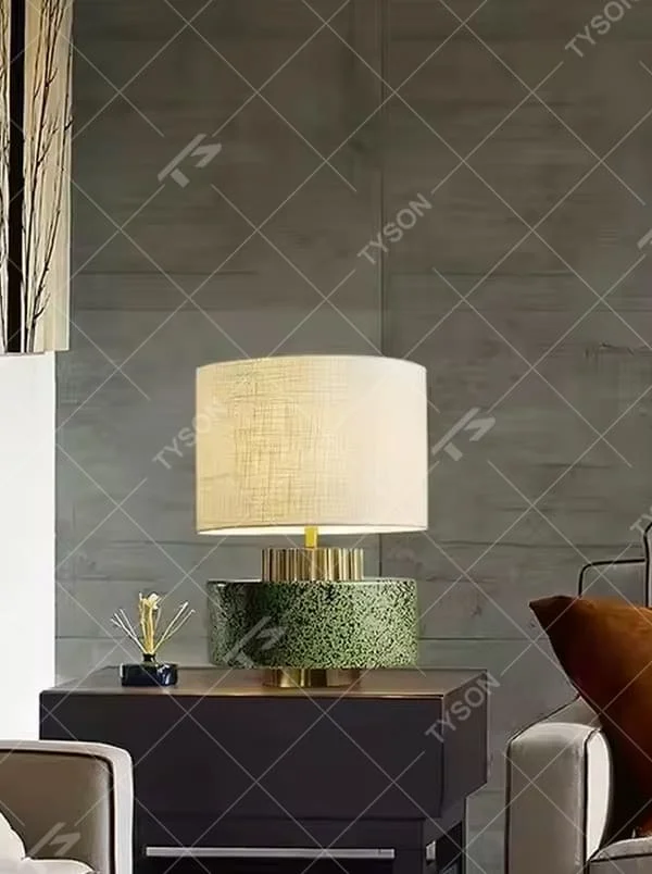 This is a modern artistic green and gold table lamp, with a green textured base, golden metal ring, and beige fabric circular lampshade, featuring a minimalist and grand design with both artistic sense and texture. It is used for local desktop lighting and decoration, creating a modern artistic atmosphere. It is suitable for modern minimalist home bedroom bedside tables, artistic-style living room side tables, high-end hotel guest rooms and other places.