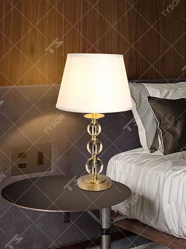 This is a luxury crystal ball table lamp with a golden metal frame, multi-layer transparent crystal balls, and a beige fabric lampshade, featuring a minimalist luxury design. It is used for local desktop lighting and decoration, creating a warm and luxurious ambiance. It is suitable for luxury-style home bedroom bedside tables, high-end hotel guest rooms, modern soft decoration display spaces and other places.