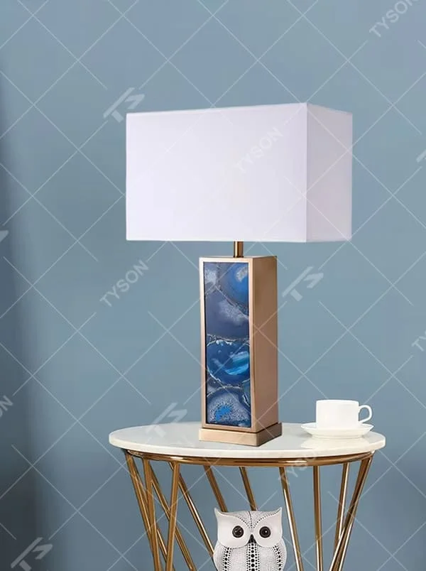 This is a luxury artistic agate-textured table lamp, with a golden metal frame inlaid with blue agate texture decoration and a white square fabric lampshade, featuring a minimalist and grand design with both artistic sense and luxury texture. It is used for local desktop lighting and decoration, creating a luxury artistic atmosphere. It is suitable for luxury-style home bedroom bedside tables, high-end hotel guest rooms, art exhibition hall display tables and other places.
