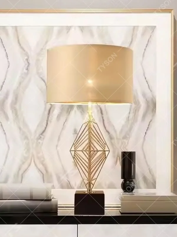 This is a luxury geometric diamond-shaped table lamp with a golden geometric hollow diamond bracket and a beige fabric lampshade. It features a simple and elegant design with excellent texture. It is used for local desktop lighting and decoration, creating a luxurious and warm ambiance. It is suitable for luxury-style home bedroom bedside tables, high-end hotel guest rooms, modern soft decoration display spaces and other places.