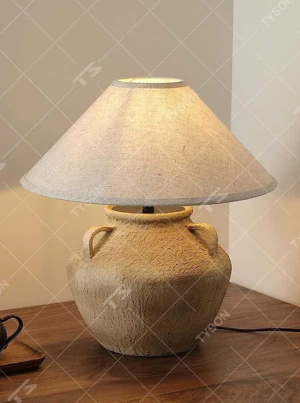 This is a retro clay pot - shaped table lamp with a beige textured clay pot base (with double handles) and a beige fabric slant lampshade, featuring a natural and rustic design with retro artistic sense. It is used for local desktop lighting and decoration, creating a warm retro atmosphere. It is suitable for wabi - sabi style home living rooms, retro - style homestays, art soft decoration display spaces and other places.