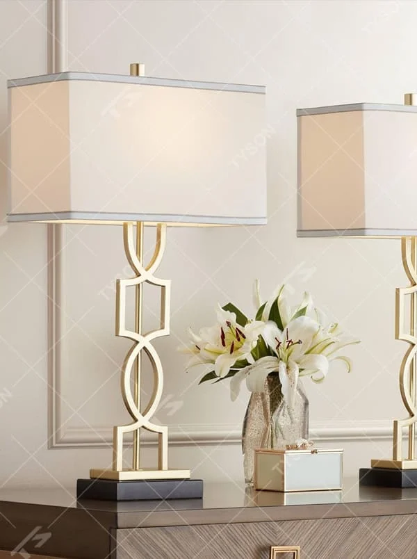 This is a luxury-style table lamp with a golden geometric hollow bracket and a white fabric square lampshade. It features a simple and elegant design with excellent texture. It is used for local desktop lighting and decoration, creating a luxurious and warm ambiance. It is suitable for luxury-style home bedroom bedside tables, high-end hotel guest rooms, modern office spaces and other places.