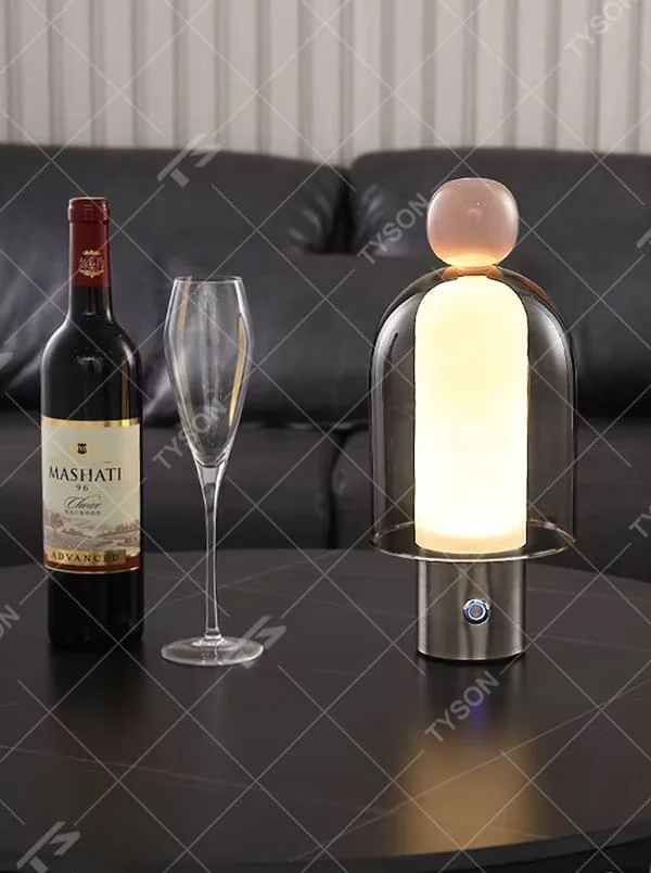 This is an artistic doll-style glass and metal table lamp, with a transparent glass shell, pink top, and metal base. Its unique shape is like an artistic doll, featuring a strong modern artistic sense. It is used for local desktop lighting and decoration, creating an artistic and modern atmosphere. Suitable for art exhibition halls, corners of modern home studies, designer studios and other places.