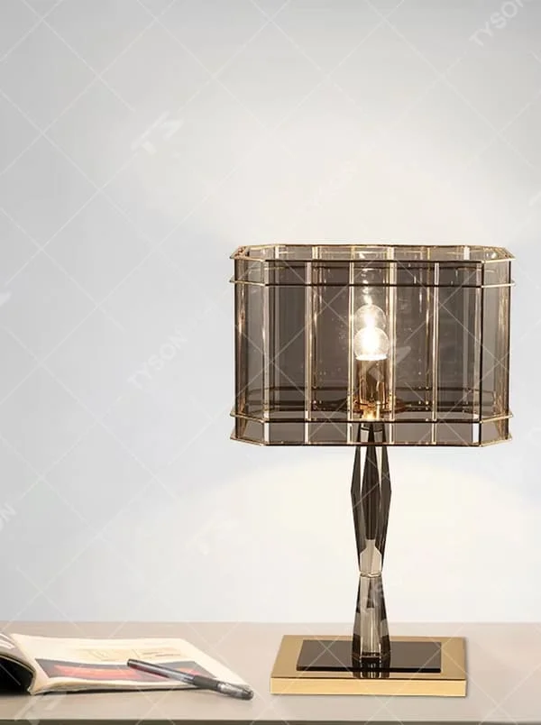 This is a light luxury and luxurious smoky gray glass metal table lamp, with a smoky gray glass lampshade paired with a golden metal frame, a golden base, and a geometrically cut lamp post. It features an exquisite and grand design, with soft and textured lighting through the glass. It is used for local desktop lighting and decoration, creating a light luxury and luxurious atmosphere. Suitable for light luxury-style bedroom bedside tables, luxury living room side tables, high-end hotel guest rooms and other places.