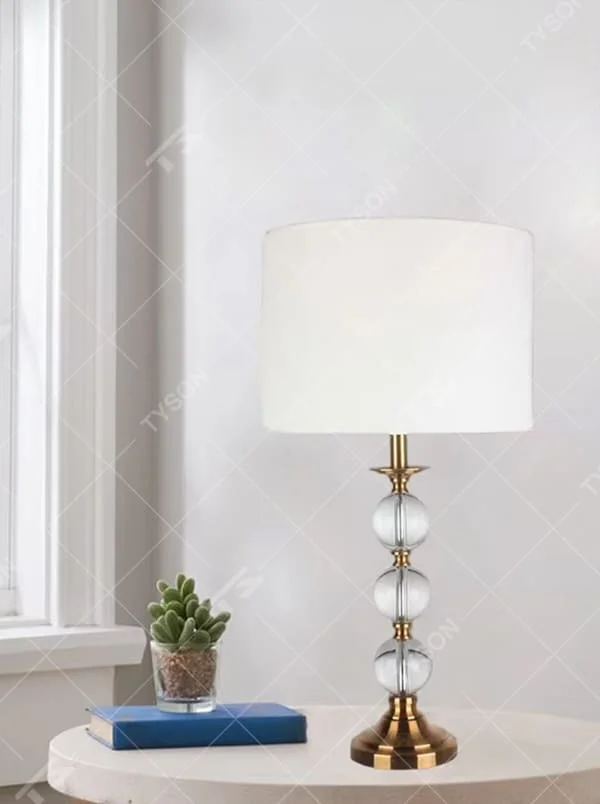 This is a light luxury exquisite crystal ball column table lamp, with a golden metal base and connectors, paired with transparent crystal balls and a white fabric round lampshade. It features a light luxury and exquisite design, with warm and soft lighting. It is used for local desktop lighting and decoration, creating a light luxury elegant atmosphere. Suitable for light luxury-style bedroom bedside tables, modern minimalist living room side tables, high-end hotel guest rooms and other places.