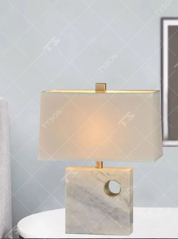 This is a light luxury elegant marble-based fabric table lamp, with a white marble base with golden ring decoration, paired with a beige fabric square lampshade and golden metal connectors. It features a minimalist light luxury design, with warm and soft lighting. It is used for local desktop lighting and decoration, creating a light luxury elegant atmosphere. Suitable for light luxury-style bedroom bedside tables, modern minimalist studies, high-end hotel guest rooms and other places.