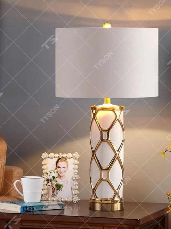 This is a luxury-style gold and white geometric hollow table lamp, with a gold geometric hollow base, white cylinder, and beige fabric circular lampshade, featuring an exquisite and elegant design with strong luxury texture. It is used for local desktop lighting and decoration, creating a luxury and warm atmosphere. Suitable for luxury-style home bedroom bedside tables, high-end hotel guest rooms, European-style living room side tables and other places.