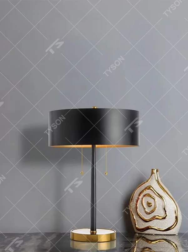 This is a light luxury high-end black and gold cylindrical table lamp, with a black silk cylindrical lampshade paired with a black lamp pole, and golden base & drawstring switch as embellishments. The design is simple, grand and high-quality, with soft and stylish lighting. It is used for local desktop lighting and decoration to create a light luxury high-end atmosphere. Suitable for light luxury-style bedroom bedside tables, modern minimalist living room side tables, high-end hotel guest rooms and other places.