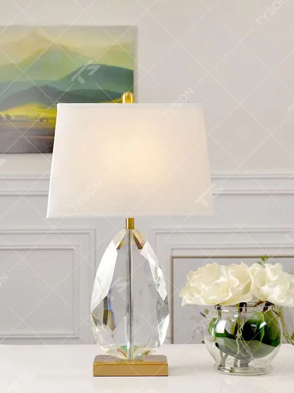 This is a light luxury exquisite water-drop shaped crystal table lamp, with a transparent cut water-drop crystal base paired with golden metal parts and a white square fabric lampshade. The design is simple and luxurious, with soft and elegant lighting. It is used for local desktop lighting and decoration, creating a light luxury exquisite atmosphere. Suitable for light luxury-style bedroom bedside tables, modern minimalist living room side tables, high-end hotel guest rooms and other places.