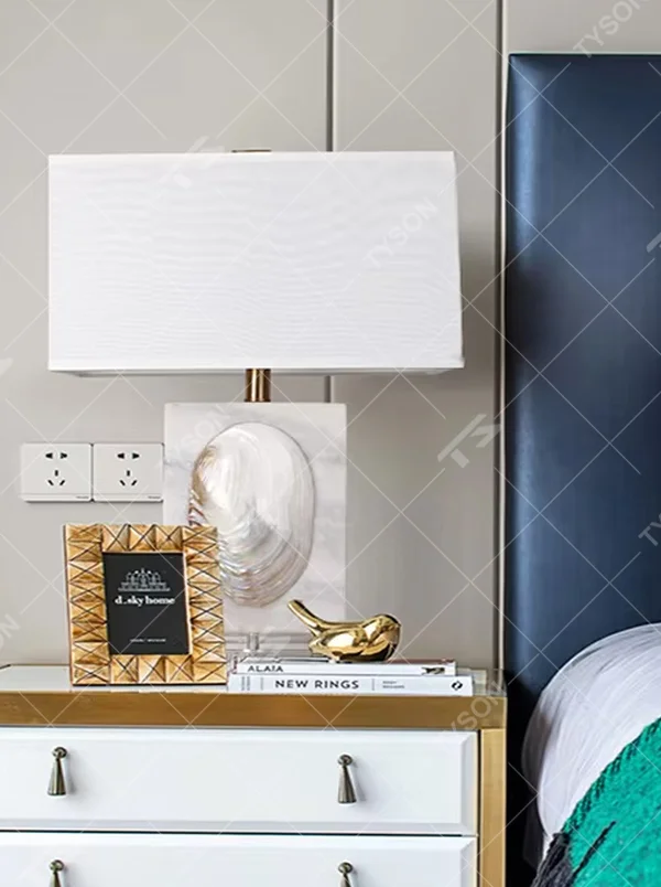 This light luxury modern table lamp features a light-colored stone base with shell relief, paired with gold metal connectors and an off-white fabric rectangular lampshade. It serves both lighting and decorative purposes, ideal for light luxury spaces such as bedroom bedside tables, living room side tables, and hotel guest rooms.