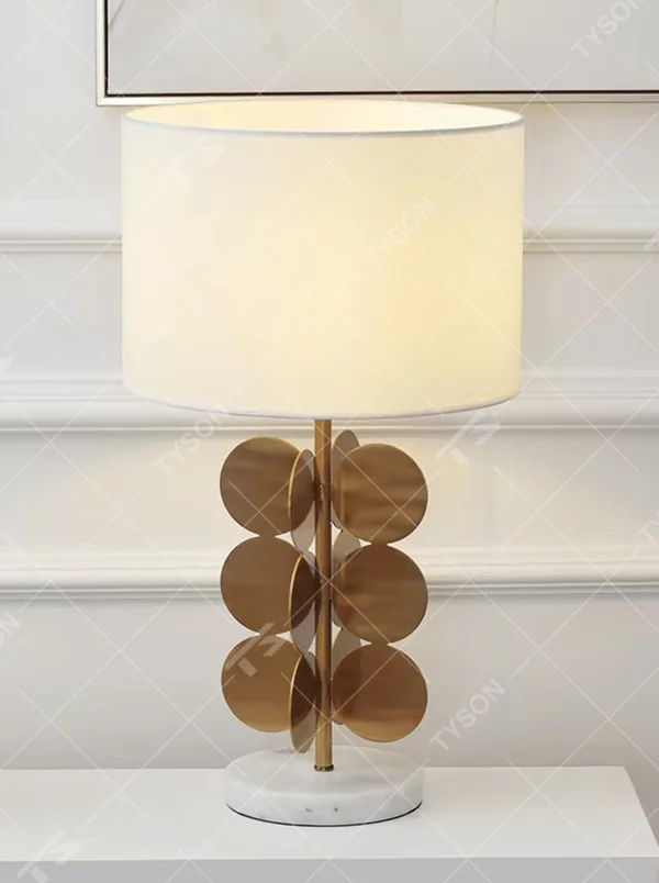 This light luxury table lamp features a white fabric cylindrical lampshade on top, a lamp post with a staggered stack of gold metal discs, and a white marble base, serving both lighting and decorative purposes, ideal for living room side tables, bedroom bedside tables, light luxury homestays and other spaces.
