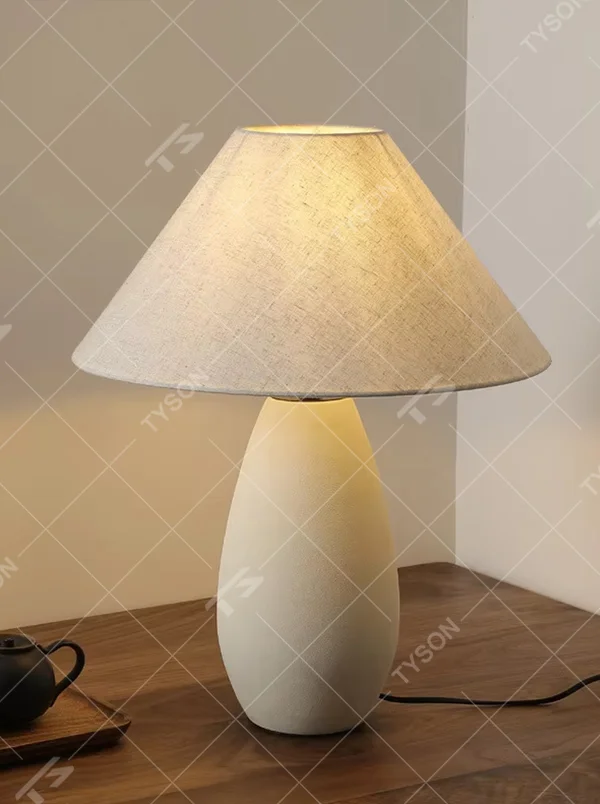 This is a wabi-sabi style minimalist table lamp, featuring an off-white frosted ceramic base paired with a linen fabric lampshade. The light is soft and warm, serving both lighting and decorative purposes, suitable for scenarios pursuing a warm and relaxed feeling such as bedroom nightstands, homestay guest rooms, Japanese-style tea rooms, and wabi-sabi style living rooms.
