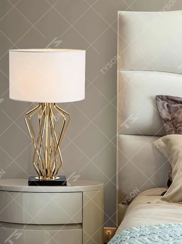 This is a light luxury artistic table lamp, featuring a gold metal geometric hollow lamp body paired with a black marble base and a white fabric lampshade. It emits soft and uniform light, serving both lighting and decorative purposes, suitable for bedroom nightstands, hotel guest rooms, light luxury living room side tables, high-end clubs and other scenarios.