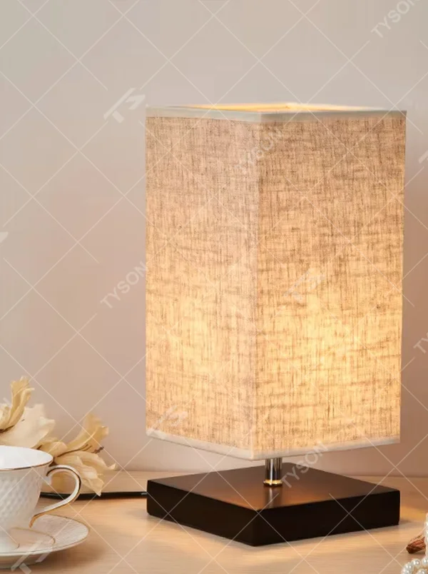 This is a minimalist square fabric table lamp, featuring a light beige linen lampshade paired with a black solid wood base. It emits soft and warm light, serving both lighting and decorative purposes, suitable for bedroom nightstands, desks, homestay guest rooms, living room side tables and other scenarios.