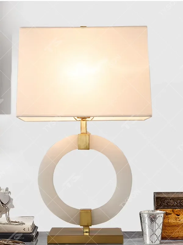 Modern light luxury style table lamp with a white natural stone/acrylic ring paired with gold metal brackets and base, and a rectangular white fabric lampshade. It emits soft and even light, combining decorativeness and practicality, suitable for lighting and decoration in spaces such as bedroom nightstands, living room side tables, and hotel guest rooms.
