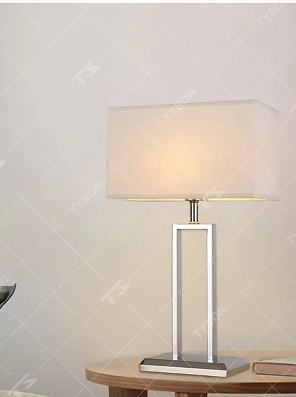 Modern minimalist table lamp with a chrome-plated metal frame and a rectangular off-white fabric lampshade, emitting soft and warm light. It is both decorative and practical, suitable for lighting and decoration in living rooms, bedrooms, studies and other spaces.