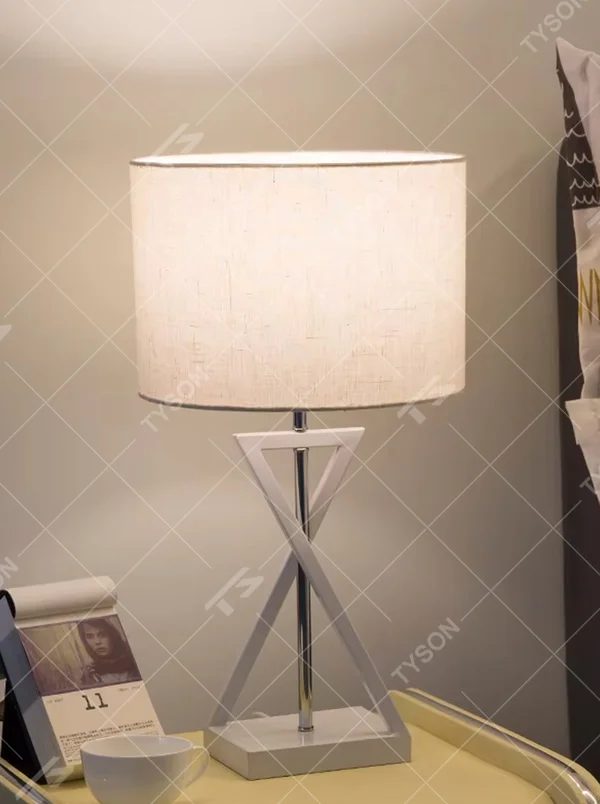 This is a modern minimalist fabric table lamp, featuring an off-white linen lampshade paired with a geometric cross-shaped white and silver dual-color lamp base, which has both lighting and decorative functions, suitable for bedroom nightstands, study desks, hotel guest rooms and other spaces.