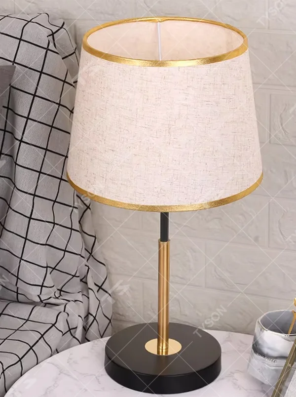 Modern light luxury fabric table lamp, with an off-white fabric lampshade (with gold trim on the top and bottom edges) paired with a black-gold spliced lamp pole and a black disc base, emitting soft and warm light, combining decorativeness and practicality, suitable for lighting and decoration in spaces such as bedroom nightstands, living room side tables, and studies.