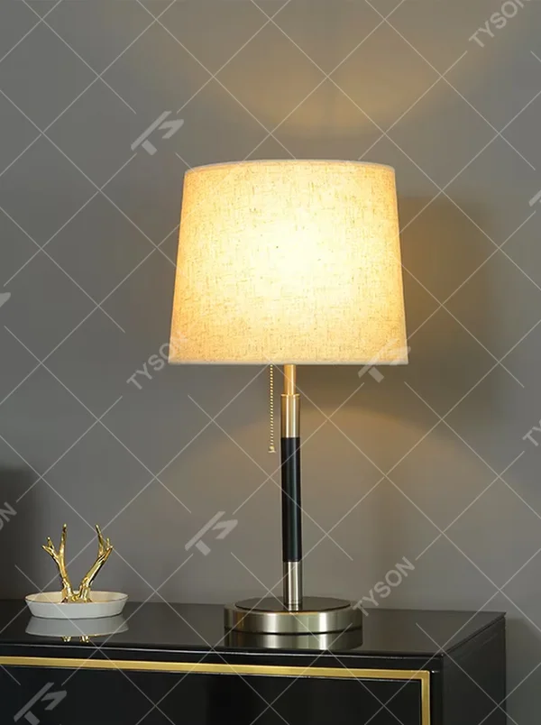 This is a minimalist light luxury table lamp, featuring an off-white linen fabric lampshade paired with a black and gold dual-color metal lamp pole, with a pull-chain switch. It emits soft and warm light, serving both lighting and decorative purposes, suitable for bedroom nightstands, living room side tables, studies, hotel guest rooms and other scenarios.