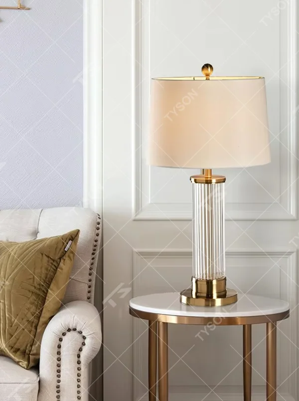 This is a light luxury crystal metal table lamp, with an off-white fabric lampshade edged with gold trim, a lamp post made of vertical striped transparent crystal paired with gold metal splicing, and a gold metal base, featuring both decorative and lighting functions, ideal for high-end indoor scenarios such as bedroom nightstands and living room side tables.