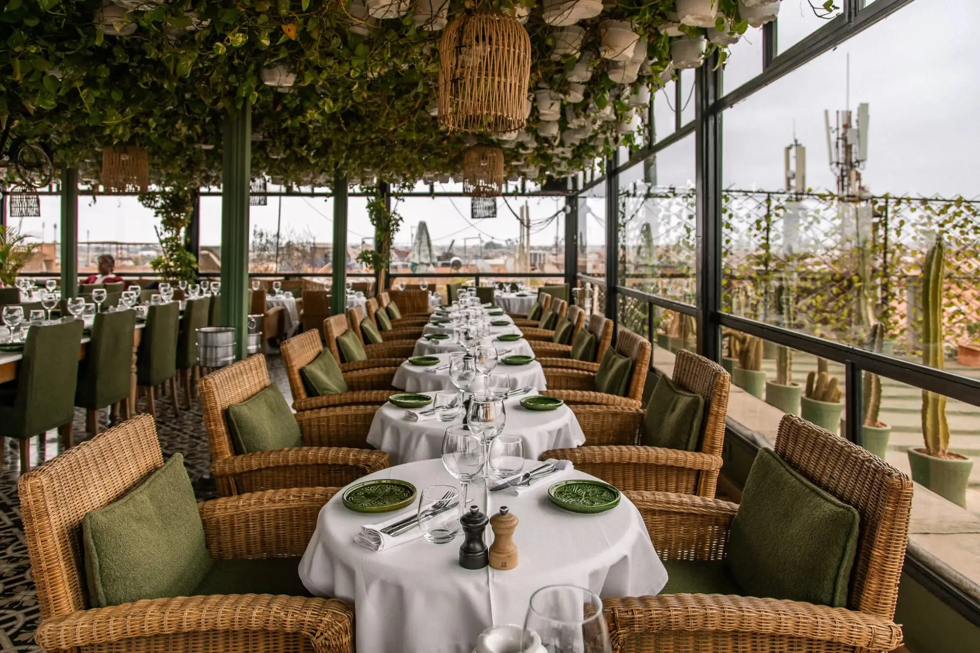 A Traditional Restaurant in Marrakech, featuring a lush, plant-covered rooftop dining area with wicker chairs, neatly set tables, and panoramic city views.