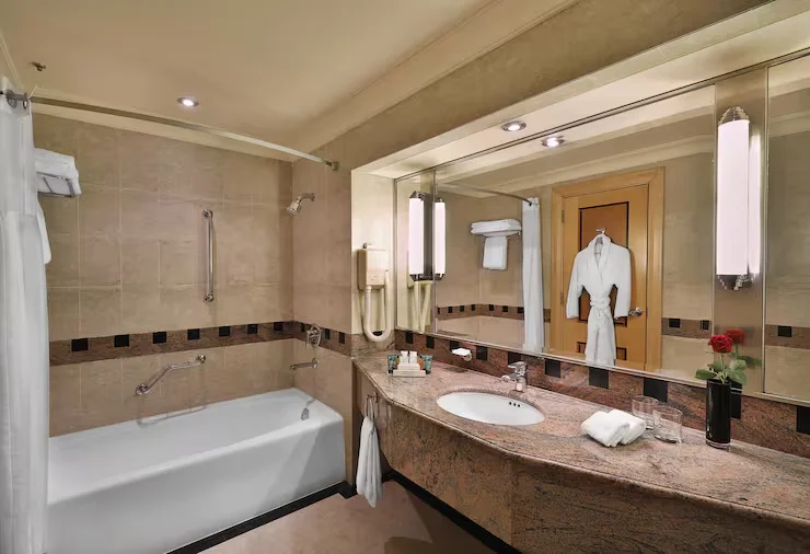 Elegant business hotel bathroom with a curved dark granite vanity, white soaking tub with grab bars, and a large framed mirror. Vertical Hotel Bathroom Mirror Lights flank the mirror, providing bright and even illumination, paired with neatly arranged bathrobes and fresh rose amenities.