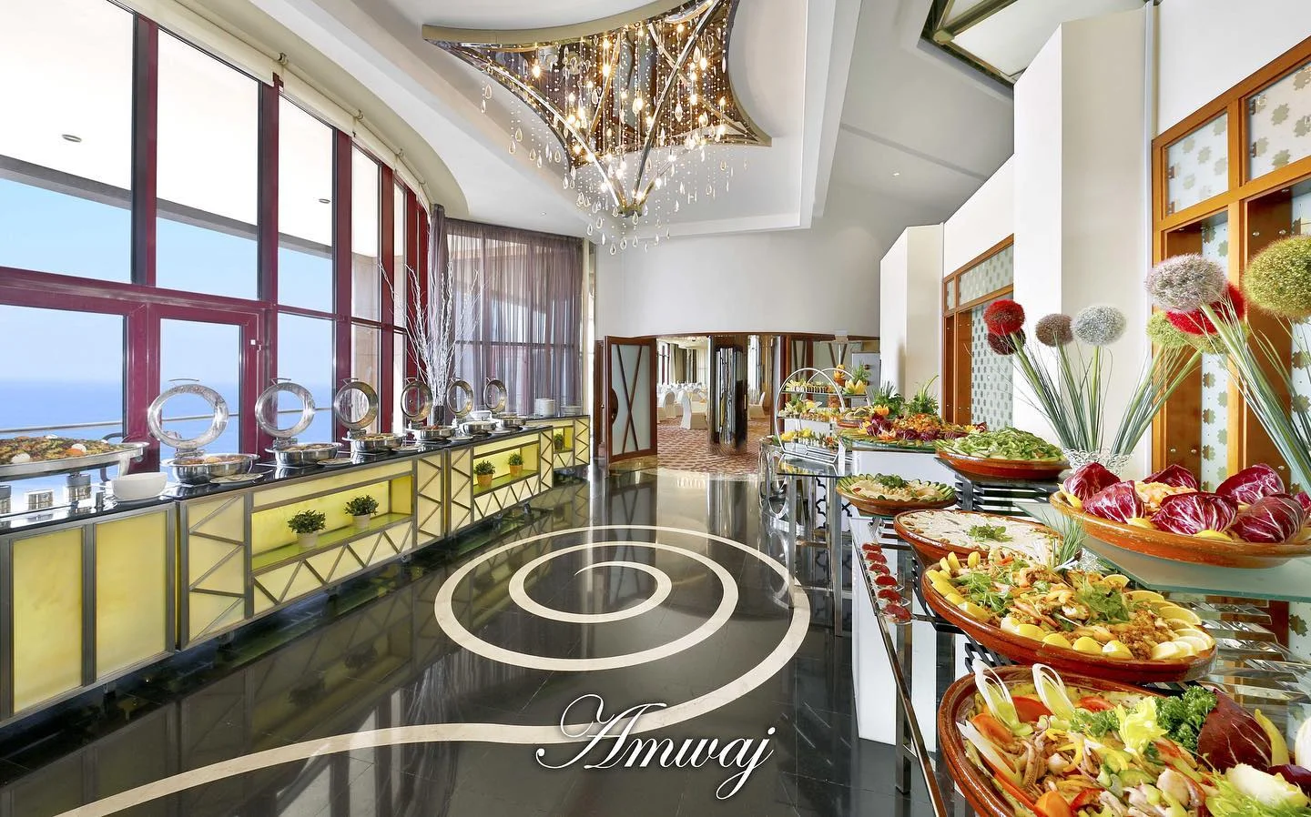 Luxury oceanfront buffet restaurant featuring a grand crystal chandelier, fresh gourmet displays, and warm accent lighting highlighting the sea view and food stations